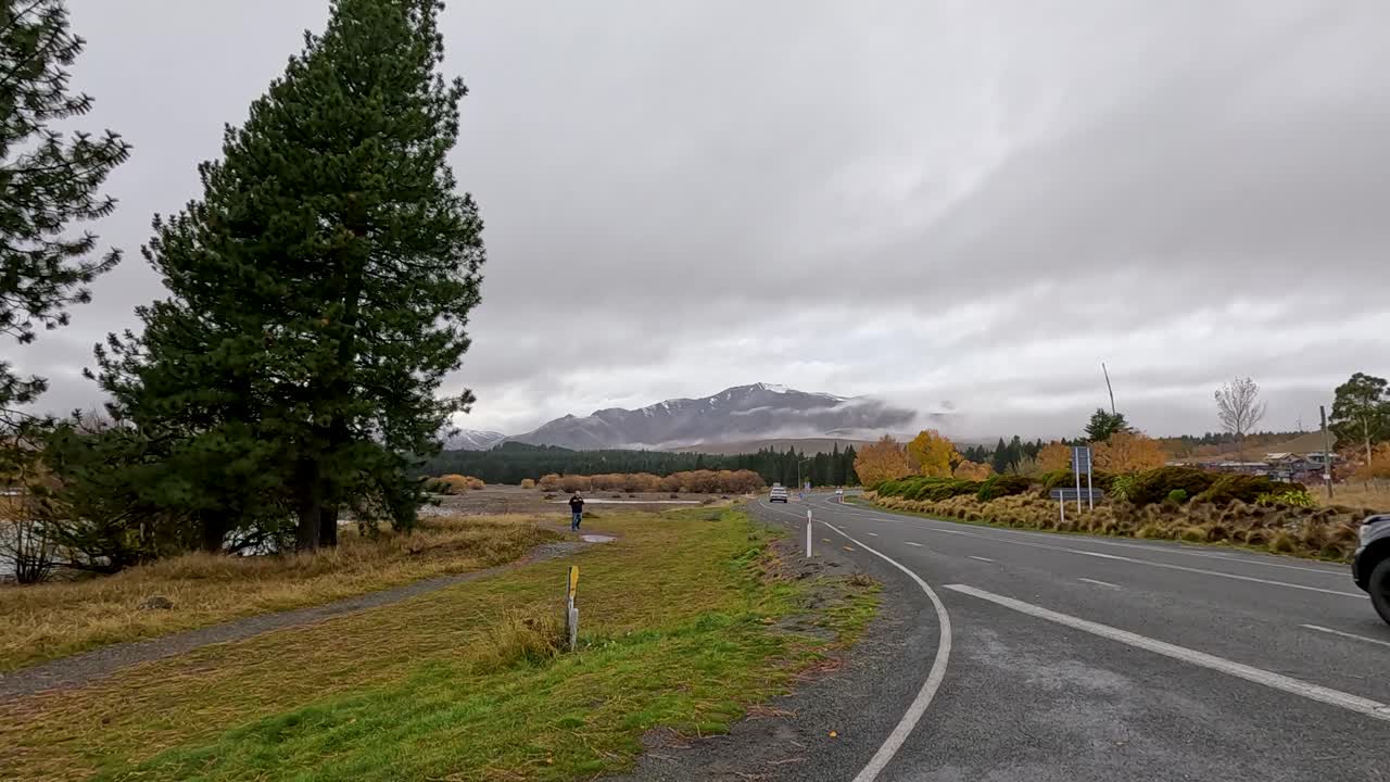 A vehicle travels along a winding lakeside road bordered by autumn trees and mountains under overcast skies, captured with a stationary wide-angle camera