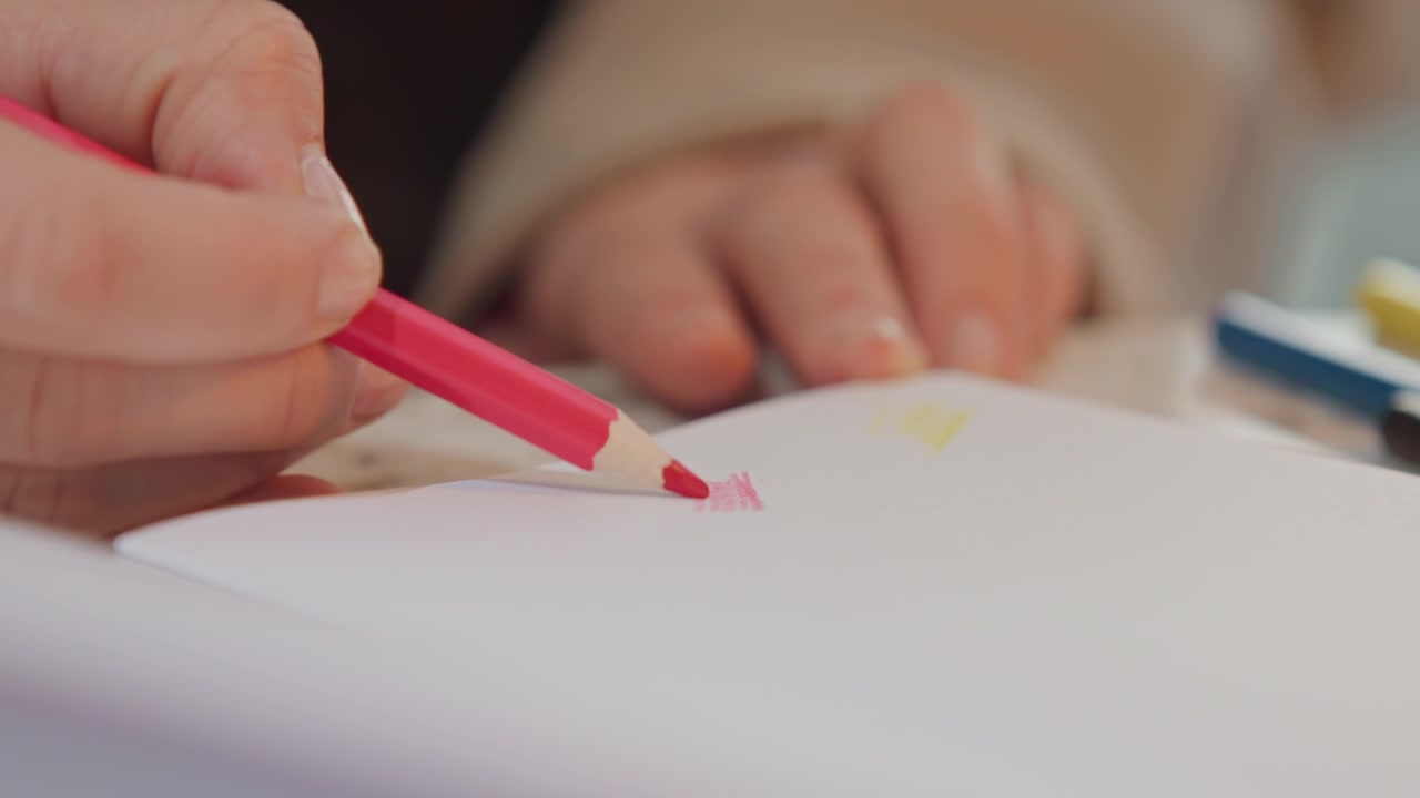 Close up of hand holding and using pink colored pencil to draw on white paper. person hands are visible with focus remaining sharp throughout as they create marks. Soft lighting highlights