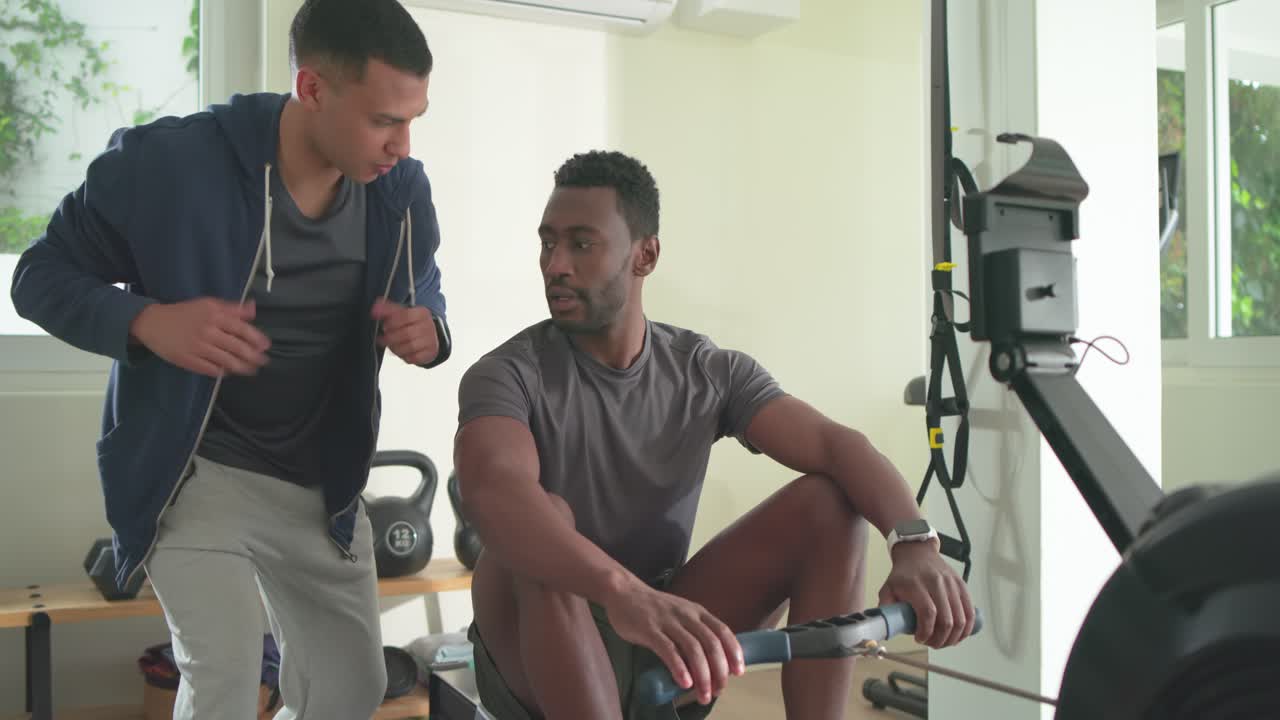 Afroamerikanischer Sportler rudert unter Anleitung des Trainers auf dem Rudergerät und verfeinert seine Form im Heim-Fitnessstudio.