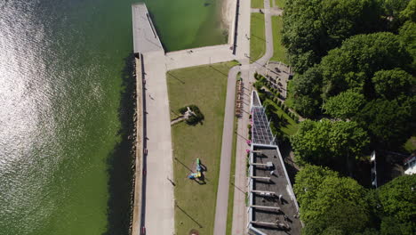 Toma-Aérea-De-Pájaros-De-Gente-Caminando-En-El-Paseo-Marítimo-De-Gdynia-Con-El-Reflejo-Del-Sol-En-El-Agua