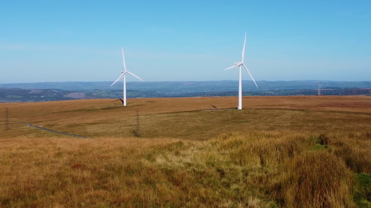 Low Angle Aerial View of Renewable Energy Wind Turbine Farm with Electricity Pylons and Countryside Road 4K and Blue Sky Background 4K