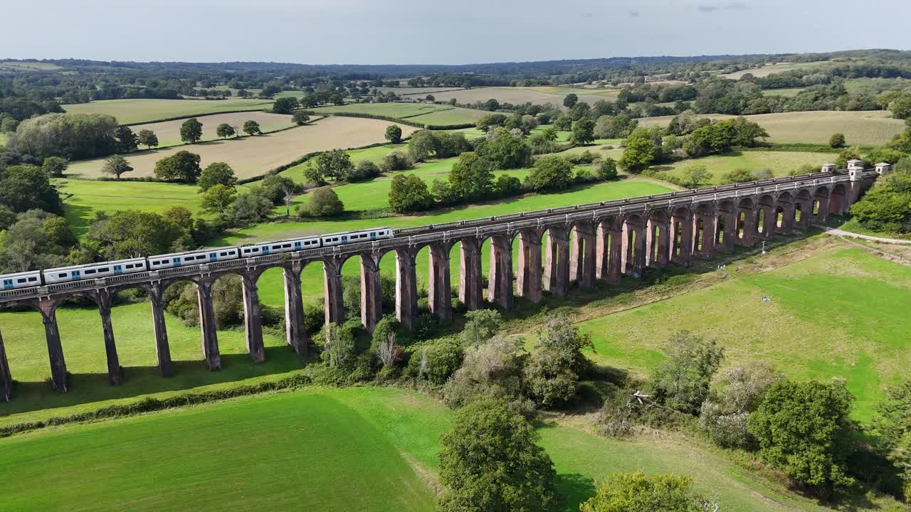 Striking drone perspective of Balcombe Viaduct, Sussex. Train moves across red-brick arches amid rolling hills, showcasing Victorian design in tranquil English landscape
