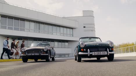 group of friends by vintage cars in front of a modern building