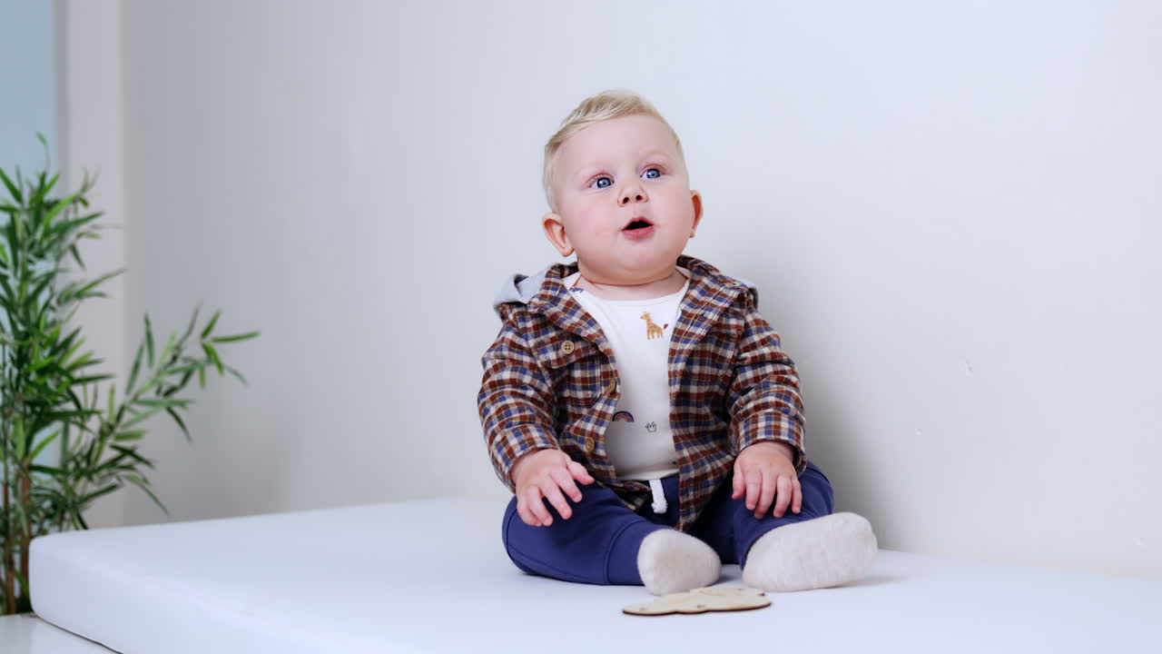 Cute blond baby boy in checkered shirt holding a wooden piece in mouth. Kid smiles adorably and pulls him by the sock.
