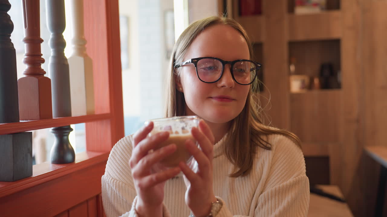 happy woman in white sweater holding coffee cup close to face while smiling gently at camera, seated indoors with warm wooden decor and blurred background