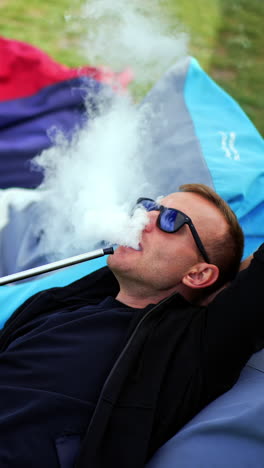 Having good rest outdoors smoking hookah. Mid-aged man in black casual clothes lies in bean bag chair and smokes shisha. Vertical video