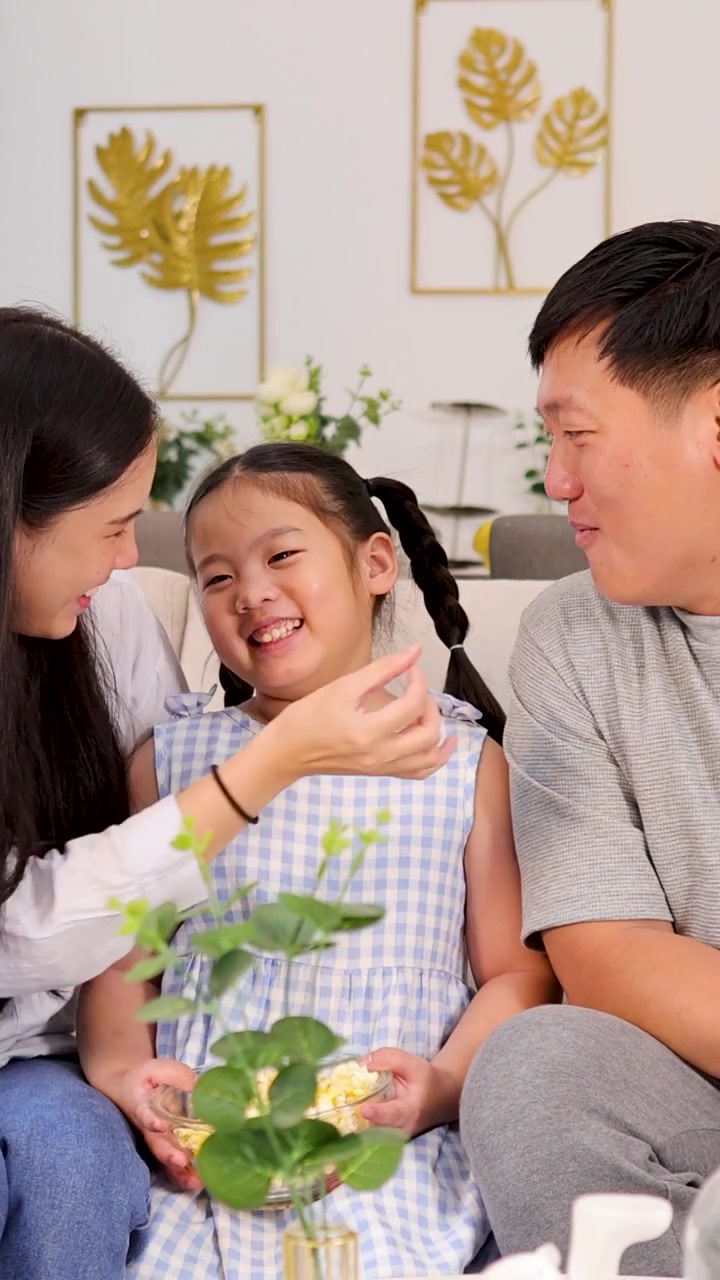 ครอบครัวชาวเอเชียที่มีความสุขกำลังเพลิดเพลินกับป๊อปคอร์นและมีปฏิสัมพันธ์ที่สนุกสนานขณะนั่งอยู่ด้วยกันบนโซฟา