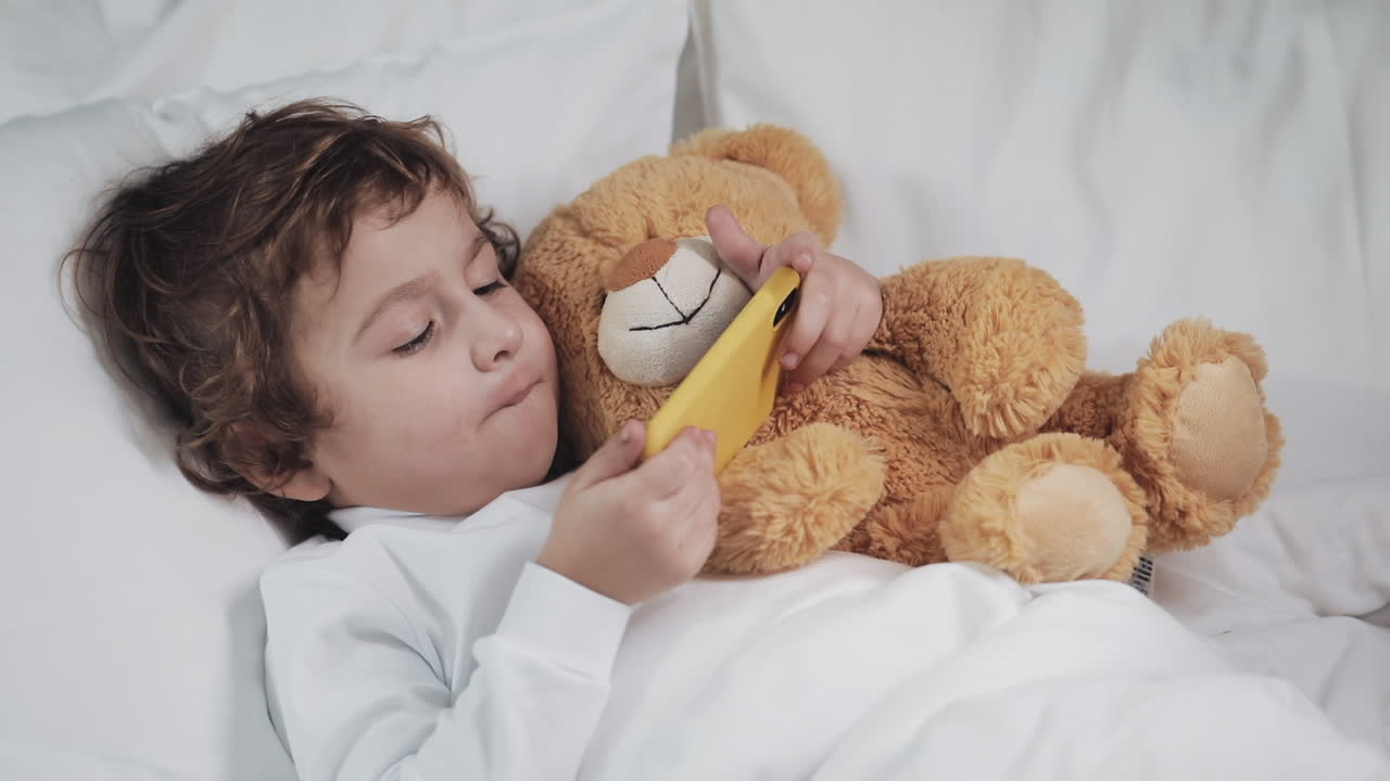 Boy playing with mobile phone in bed with teddy bear