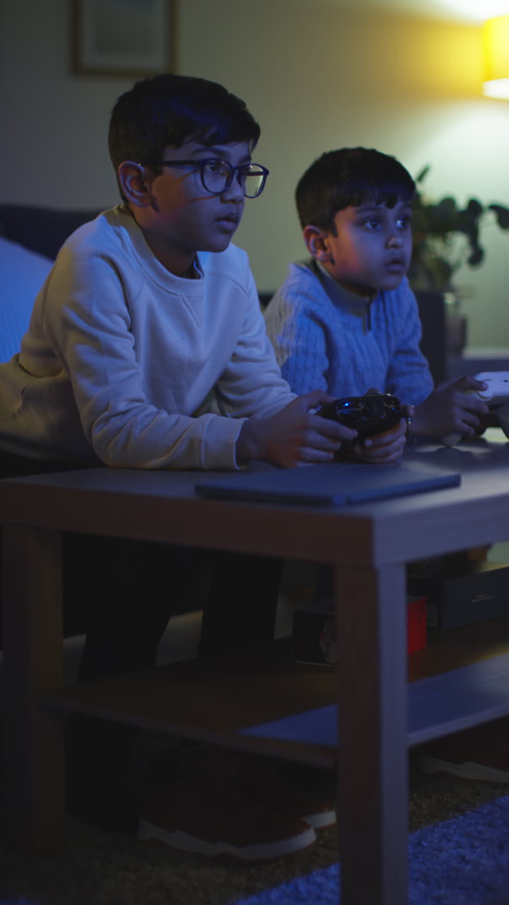 Vertical Video Of Two Young Boys At Home Playing With Computer Games Console On TV Holding Controllers Late At Night