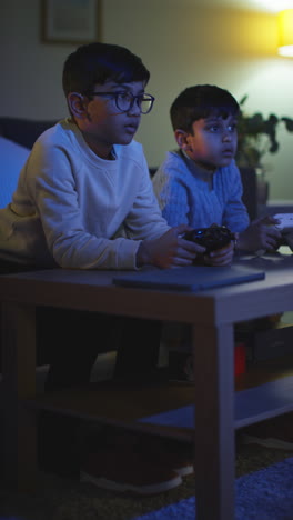 Vertikales-Video-Von-Zwei-Kleinen-Jungen,-Die-Zu-Hause-Spät-In-Der-Nacht-Mit-Einer-Computerspielkonsole-Im-Fernsehen-Spielen-Und-Controller-In-Der-Hand-Halten