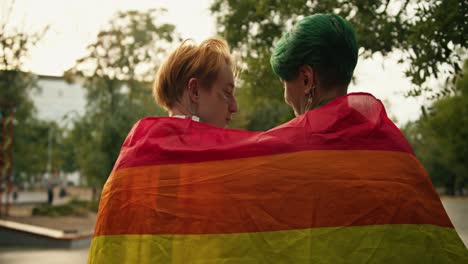 two lesbian girls sit on a fence, covered with an lgbt flag, and communicate with each other in a skate park in the summer. a girl with green short hair and a girl with blonde short hair are talking on their date at the skate park