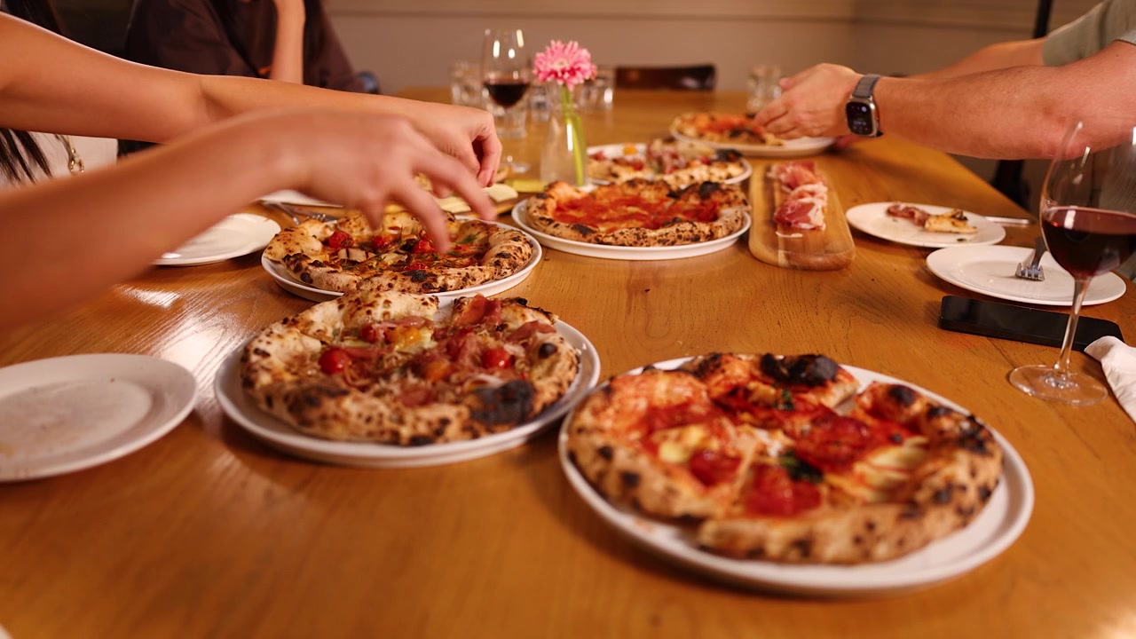 Grupo diverso comparte varias pizzas gourmet y vino tinto en una cálida mesa de comedor de madera