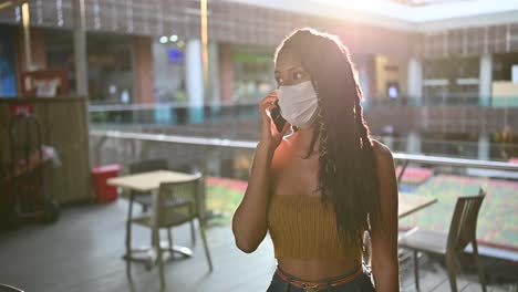 joven negra con máscara facial habla por teléfono inteligente en un centro comercial