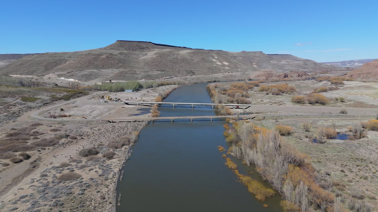 Drone ascends over Collon Cura River, showcasing Puente La Rinconada Bridge, mountains background - Neuquen, Argentina