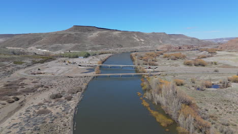 drone ascends over collon cura river, showcasing puente la rinconada bridge, mountains background - neuquen, argentina
