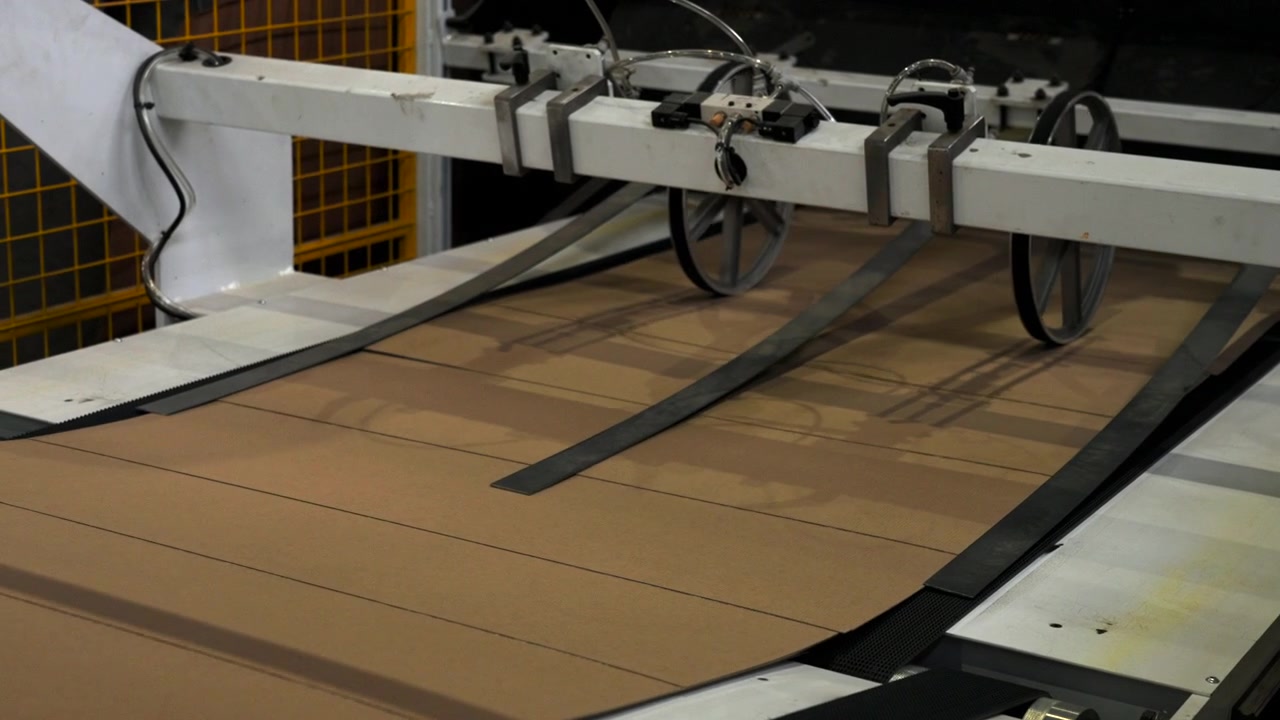 Close elevated view shows cardboard sheets moving into automated packaging equipment with metal rollers, belts, clamps, and safety mesh in dark manufacturing facility