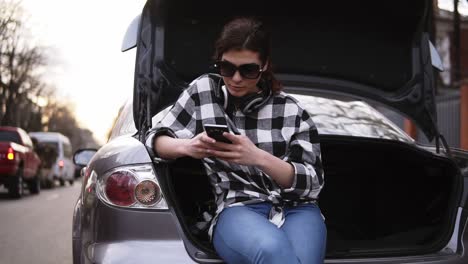 woman in sunglasses sits in open trunk of car in jeans and plaid shirt along the street. typing on mobile phone. headphones on her neck. front view