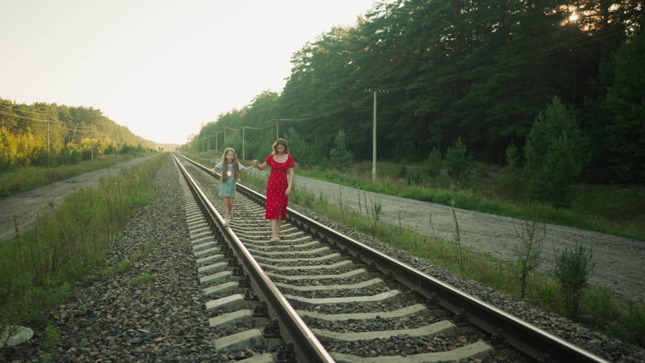 young adult in red dress walking with daughter on rail track during cool evening as child balances on beam holding mother's hand, sunlight casting soft silhouette glow over rural path and forest edge