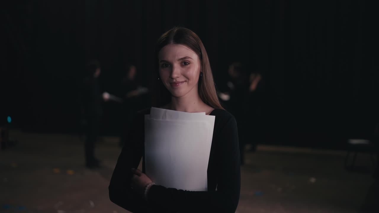 retrato de una joven actriz confiada en un traje negro que cruza los brazos en su pecho mientras sostiene papeles de guión en sus manos durante un ensayo en el teatro