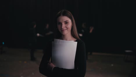 Portrait-of-a-confident-young-girl-actor-in-a-black-suit-who-crosses-her-arms-on-her-chest-while-holding-play-script-papers-in-her-hands-during-a-rehearsal-in-the-theater