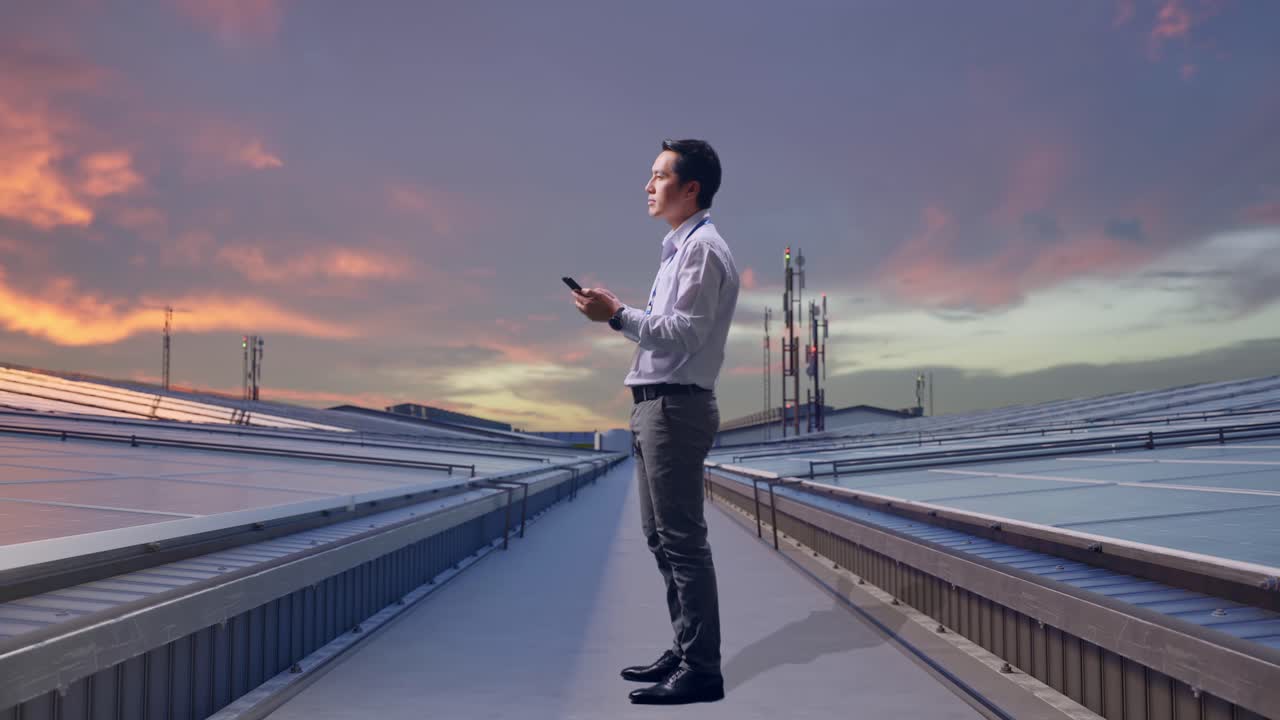 Vista laterale a figura intera di un professionista maschile asiatico in piedi con il suo telefono, mentre ispeziona pannelli solari industriali sul tetto. Osserva l'ambiente circostante prima di concentrarsi sul suo smartphone.