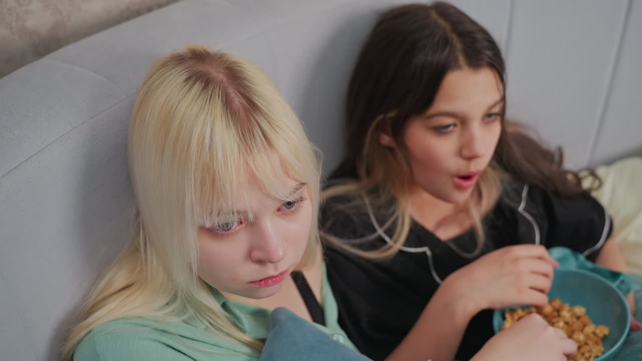 Top view of two young girls sitting closely on bed, eating popcorn from bowl and watching movie with serious expressions, surrounded by pillows and blankets in cozy indoor environment
