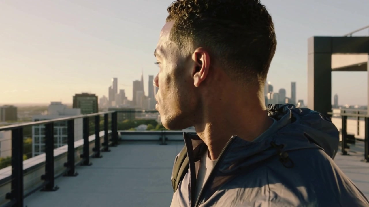 Spotting sun, turning man with pack scanning city skyline to orient on rooftop, jacket hood showing