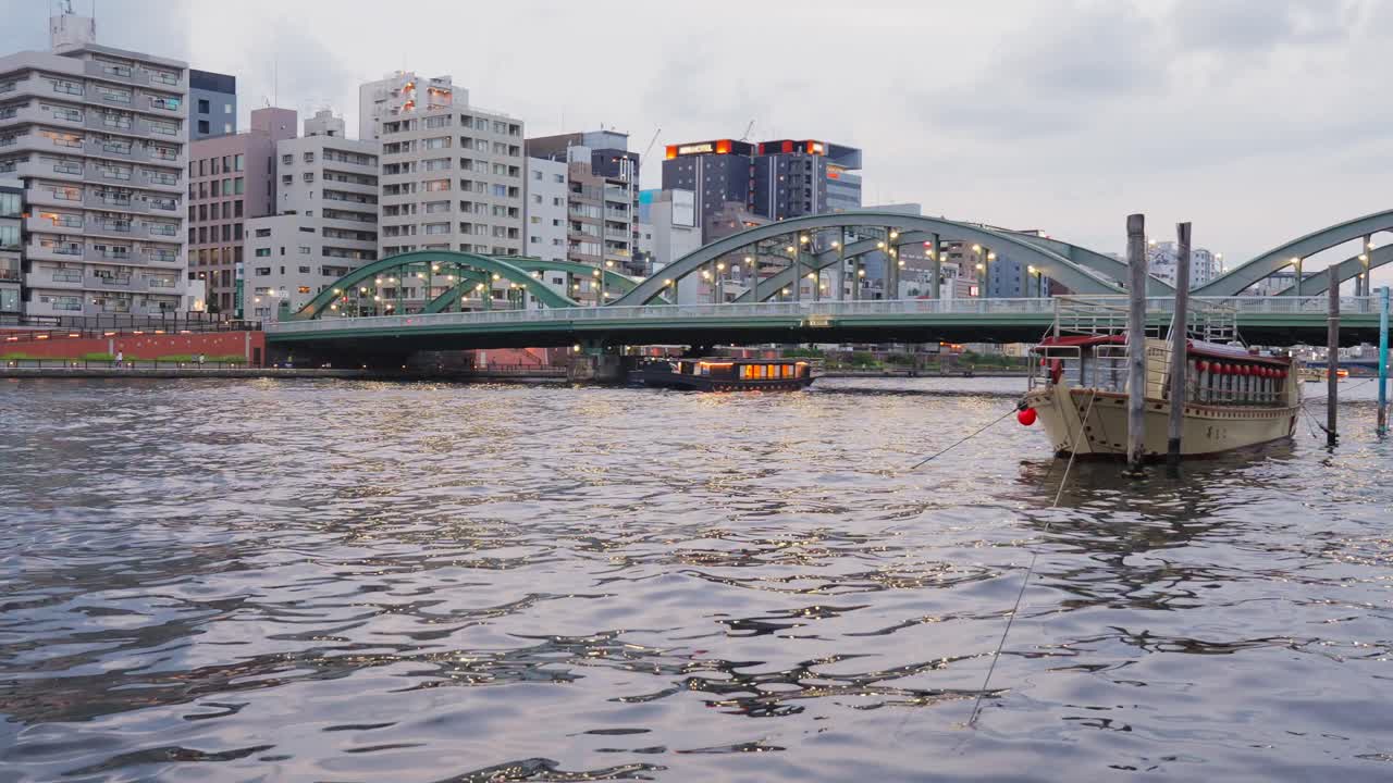 Houseboats float on the Sumida River with the lit-up Komagata Bridge visible in the background at dusk