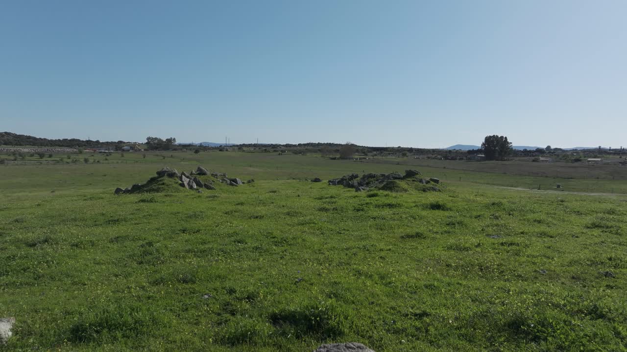 Reverse drone flight over grassy pastures and ruins of an old pigsty. With a surprise reveal, the drone passes through the Puerta de Arenas arch, uncovering its impressive stone structure.