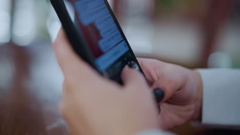 close-up of black smartphone being operated by someone with black nails, typing on screen, placed on wooden table with vibrant blurred light effect in the background