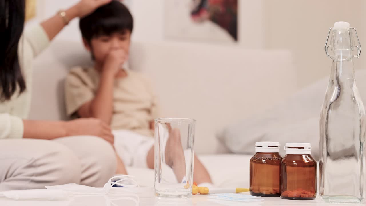Mother checks sick child's temperature on couch, surrounded by medicine bottles and comforting home setting