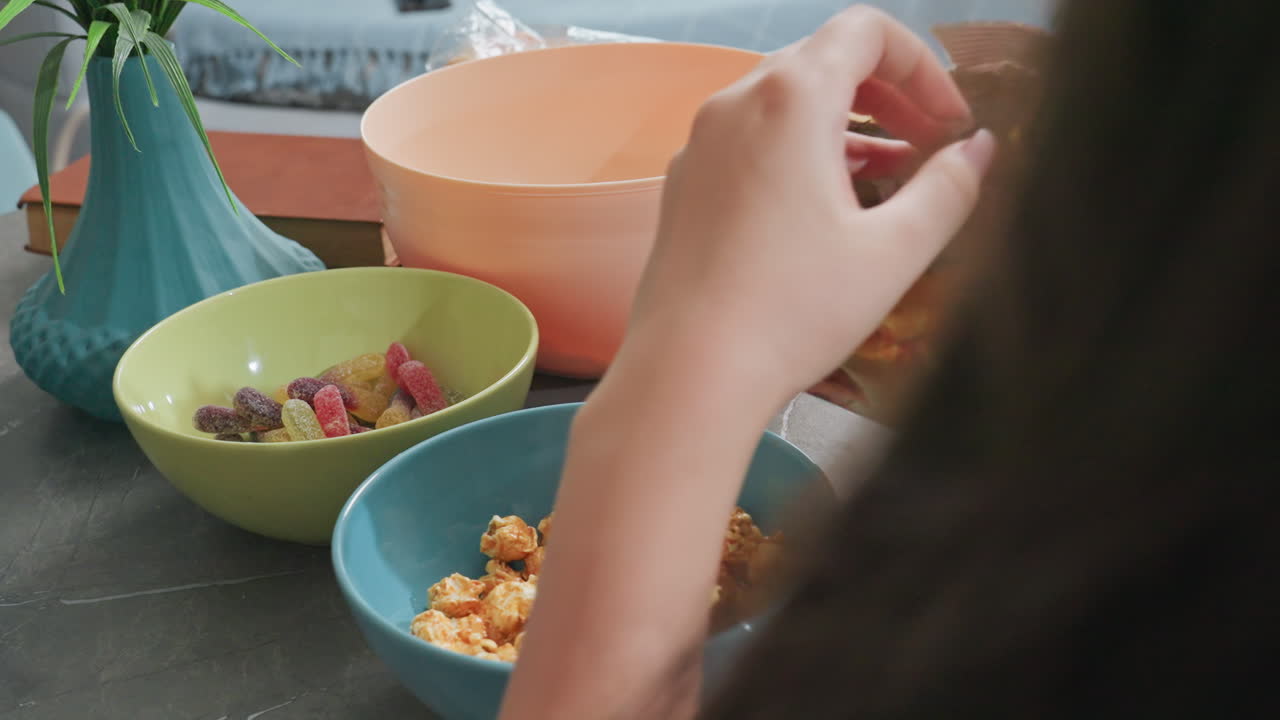 Close up rear view of woman returning caramel popcorn into packet with blue and green bowls nearby on dark table in bright indoor setting, casual snack moment with texture and detail