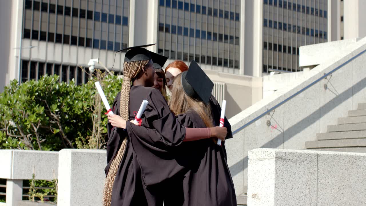 Amiche diverse in abito da cerimonia che si abbracciano tenendo in mano i diplomi e poi guardano uno smartphone in una piazza
