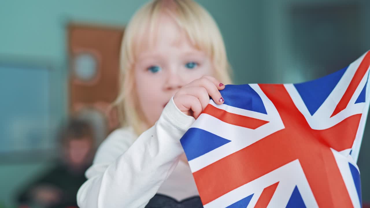 English flag in child's hands. Little girl with blond hair closes her face with the British flag. Cute child holds the flag of Great Britain. Close-up.