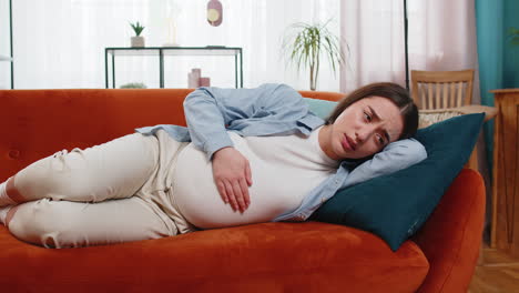 upset ill young pregnant woman feeling sick suffering from abdominal belly pain lying on home sofa