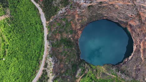 Birds-eye-view-of-blue-water-lake-formed-in-deep-sinkhole-completely-surrounded-by-tall-cliffs-adjacent-to-winding-road-in-Imotski,-Croatia