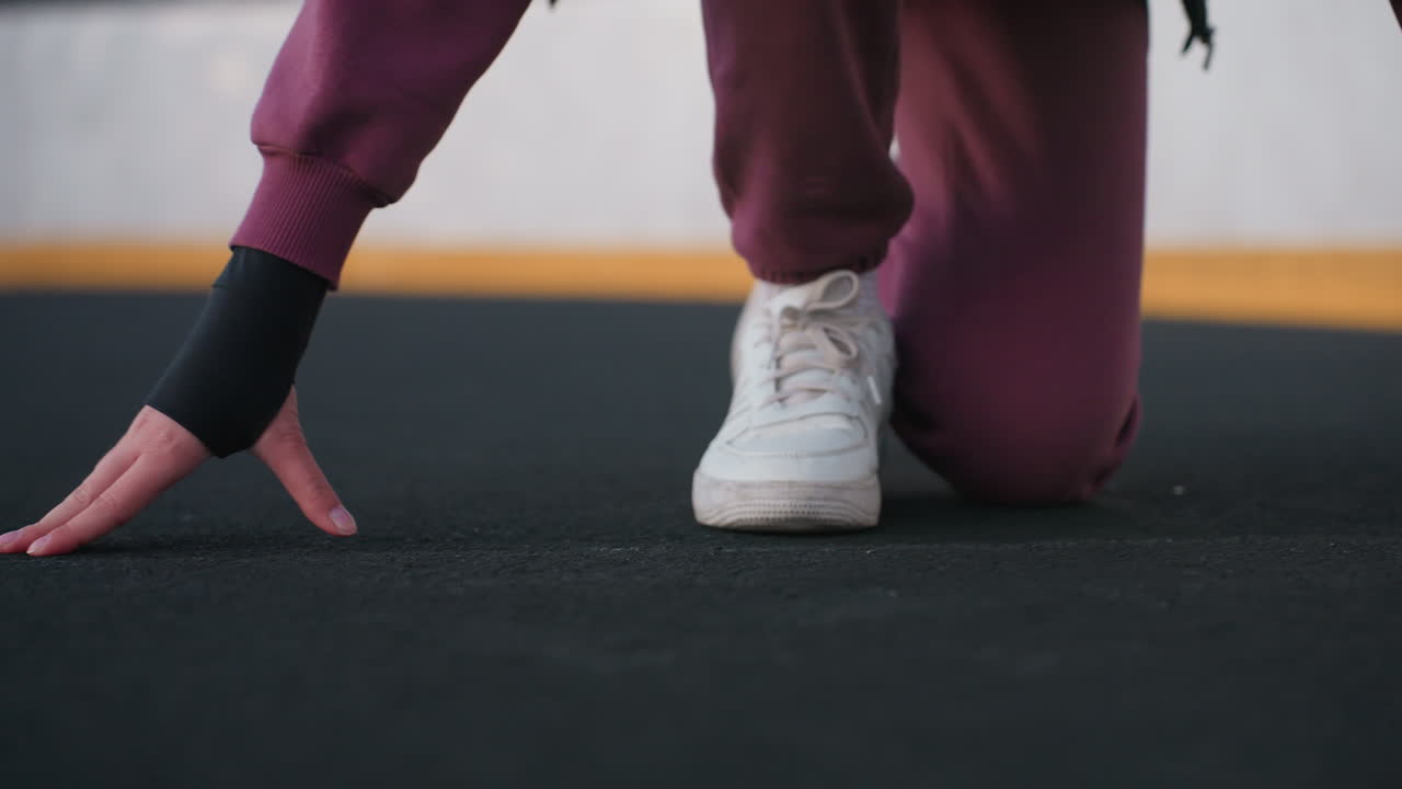 Lower view of runner placing hands on floor with one knee on ground on black asphalt sports court near white barrier topped with chain link fence wearing maroon tracksuit trousers and white sneakers