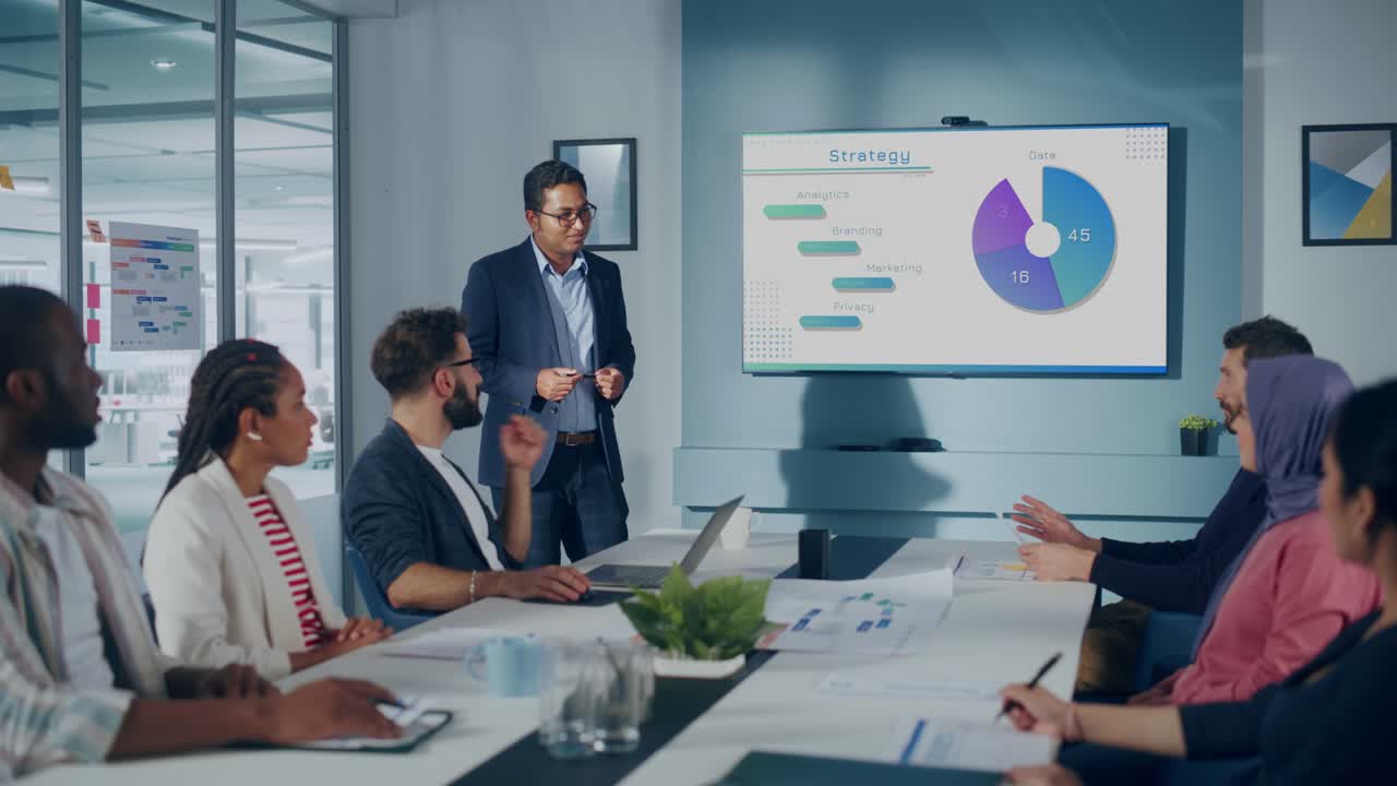 Multi-Ethnic Office Conference Room. Brilliant Indian Male CEO does Presentation for Multi-Ethnic Group of Managers Talking, Using TV Infographics, Statistics, Graphs. Innovative Businesspeople