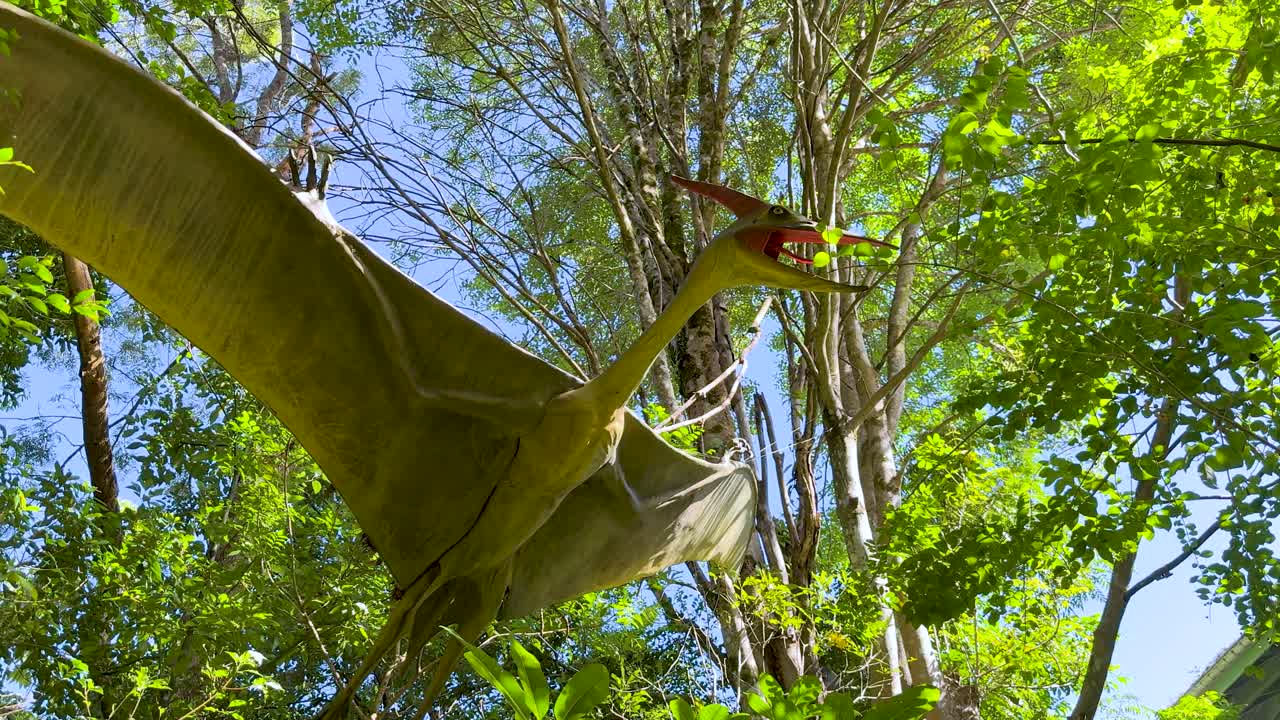 Animatronic pterodactyl flaps wings under bright daylight in a green, tree-filled park setting