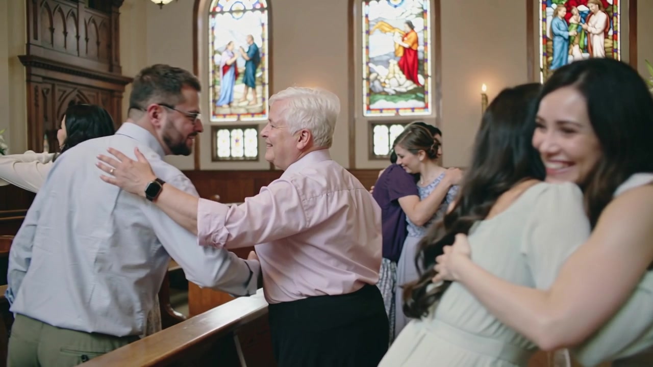 Hugging pair of women in pink sleeveless and green dresses, service ending in church, stained glass