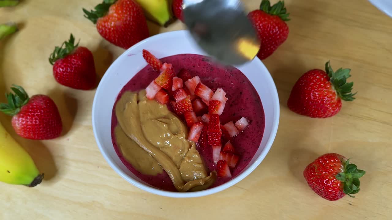 Overhead close-up of a thick acai smoothie bowl topped with creamy peanut butter and diced strawberries, surrounded by bananas—clean, appetizing b-roll for healthy recipe and wellness content