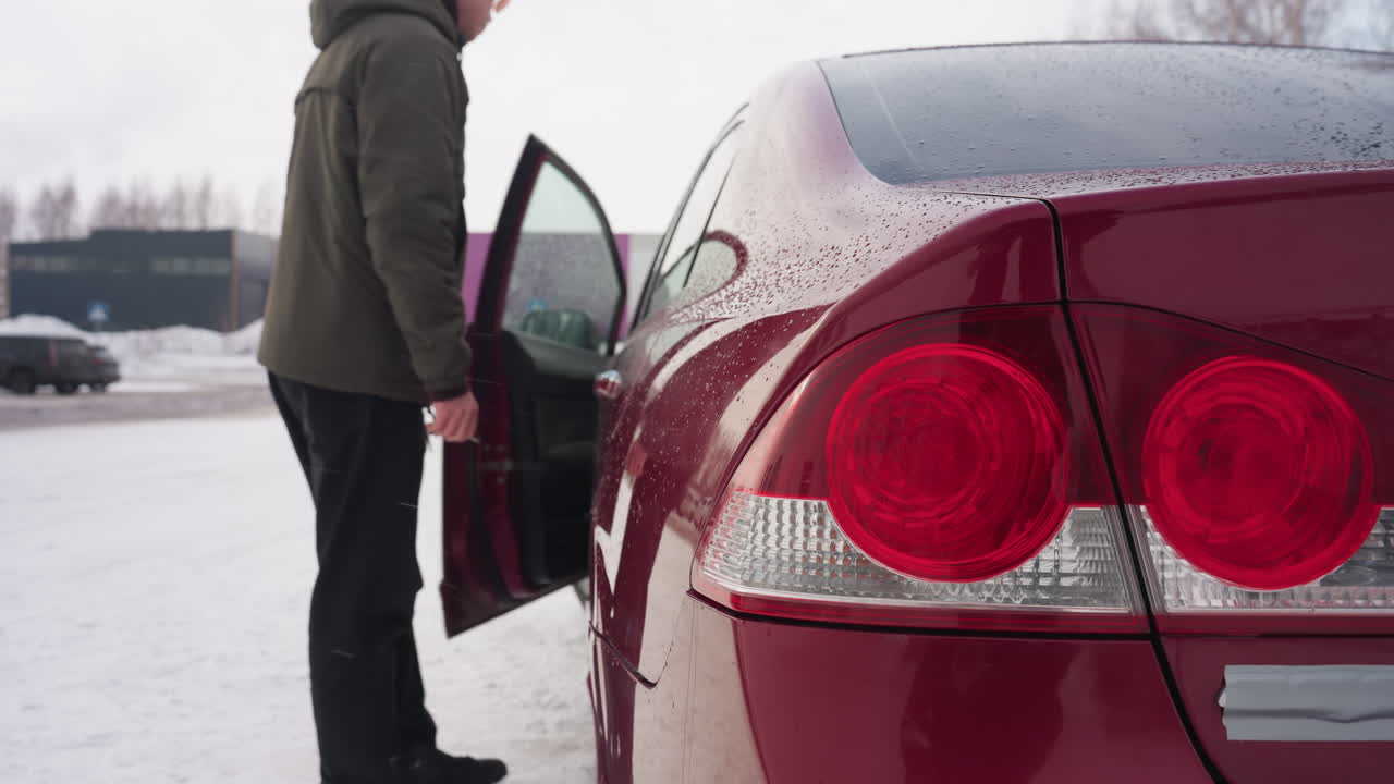 Close up of parked red car with water droplets on body and shiny rear lights, owner in winter jacket opening door and entering vehicle amidst snowy parking lot and blurred cars in background