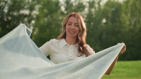 active moms swaying blanket side to side over children in sunny park, laughter and motion, green grass background, casual summer clothes conveying joyful family playtime