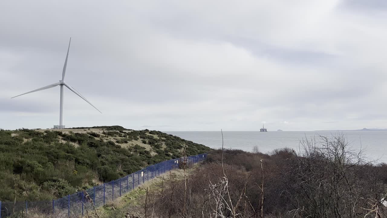 풍력 터빈이 앞면에서 빠르게 회전하는 반면, 멀리서 북해의 해안 석유 굴착장도 보인다.