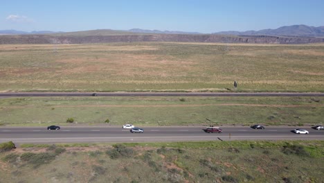 Drone-Volando-A-Lo-Largo-Del-Lado-De-La-Carretera-Que-Muestra-Tráfico-Intenso-Y-Un-Cielo-Azul-Y-Una-Pradera-En-El-Fondo