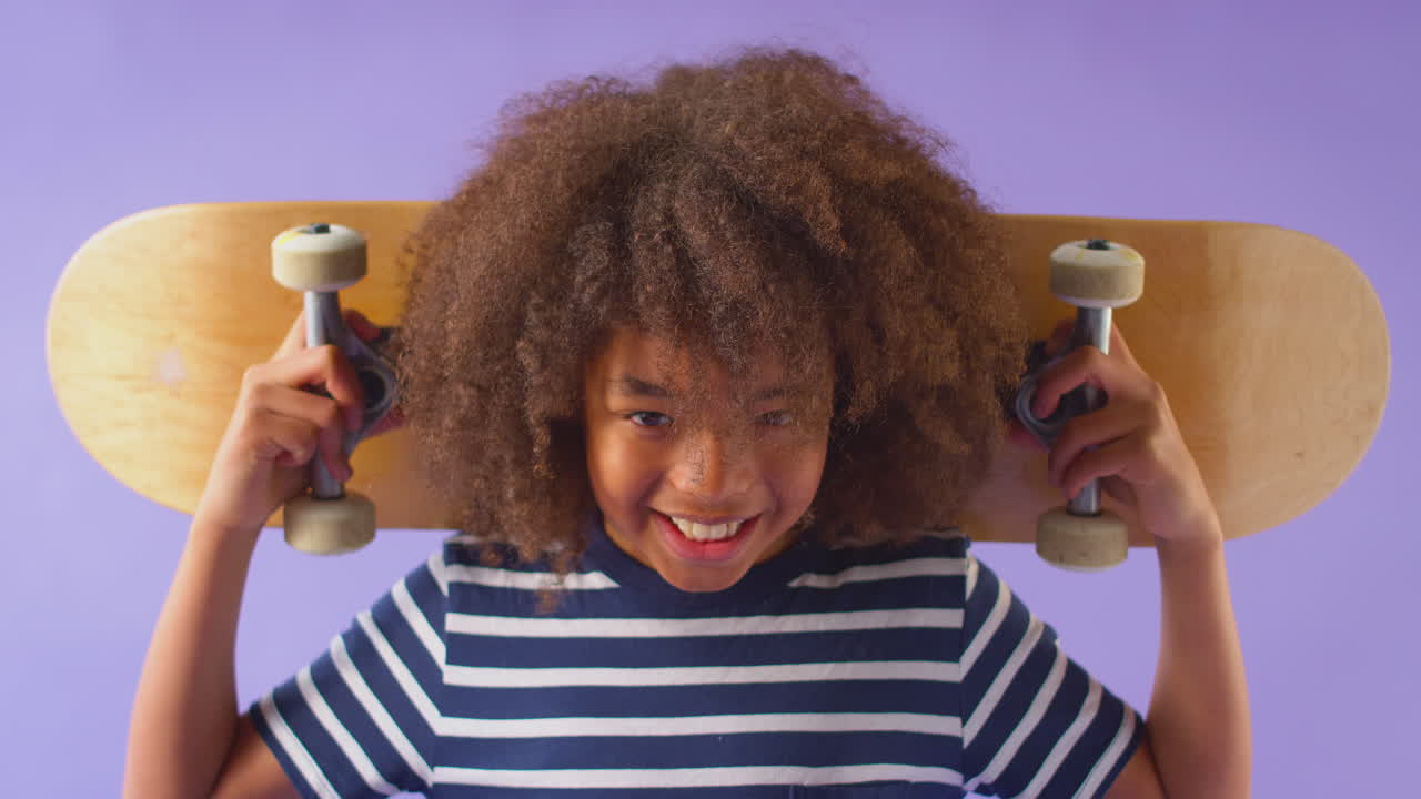 Studio Shot Of Young Boy Holding Skateboard Across Shoulders  Against Purple Background