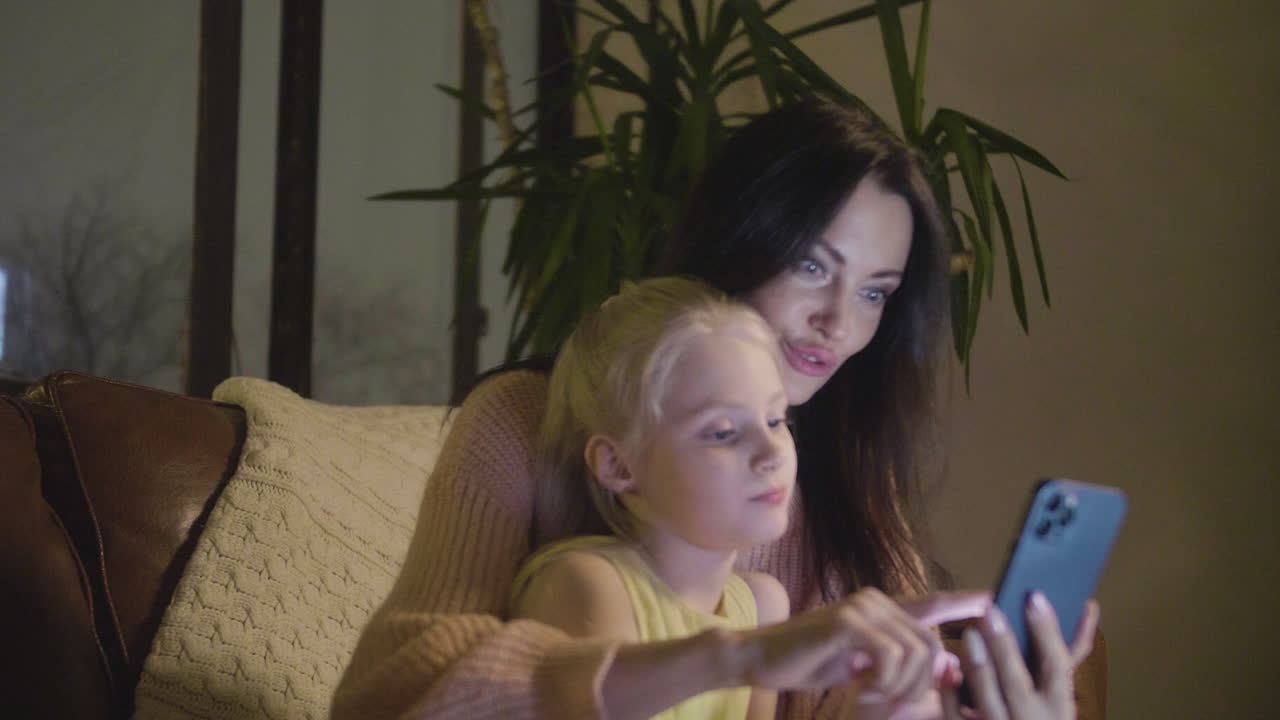 Woman Watching Something On Mobile Phone With Her Little Daughter While Sitting Together On Sofa At Home At Night 1