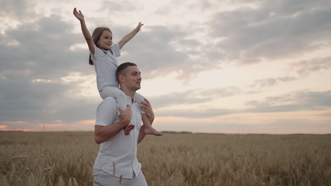 Papà porta sulle spalle la sua amata figlioletta sana sotto il sole. Al rallentatore, la bambina cammina con il padre nel campo e, libera e felice, agita le mani verso l'alto. Passeggiata nel campo.