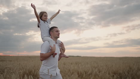 daddy carries on his shoulders his beloved little healthy daughter in sun. in slow motion the daughter walks with her father on the field and free and happy waves her hands up. walking in field.