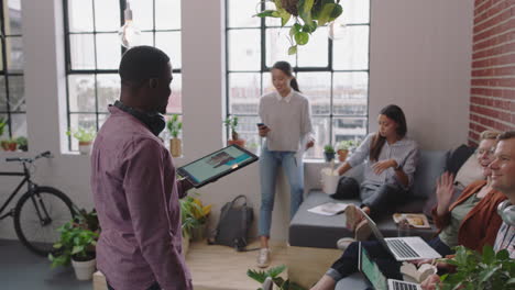 Joven-Hombre-De-Negocios-Afroamericano-Usando-Tableta-Digital-Presentando-Estrategia-De-Marketing-Líder-Del-Equipo-Hombre-Compartiendo-Ideas-Diversos-Empresarios-Colaborando-En-La-Oficina-De-La-Empresa-Emergente